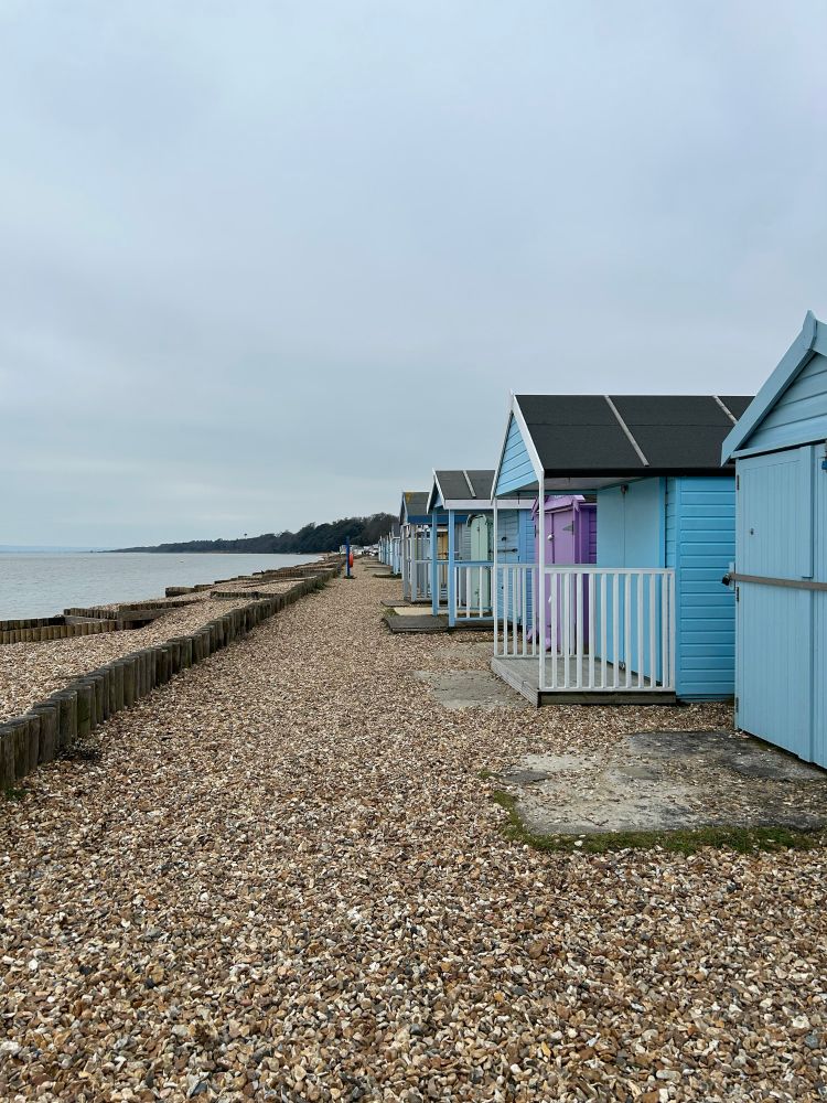 A row of beach huts lined up on the right then a pebbly beach leading to the sea.