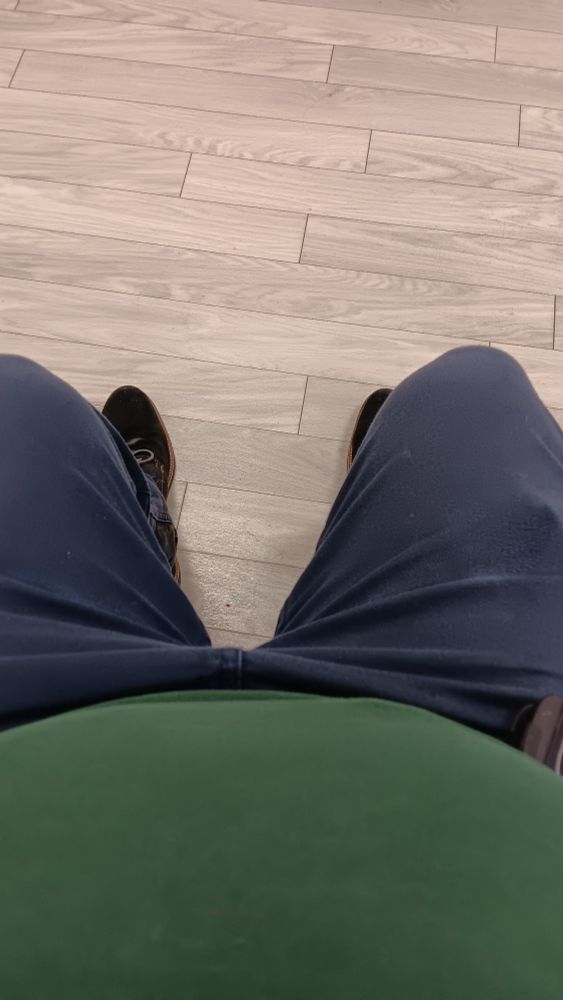 Ich sitze in blauen Jeans und grünem T-Shirt in einem Wartezimmer mit hellem Laminatboden.