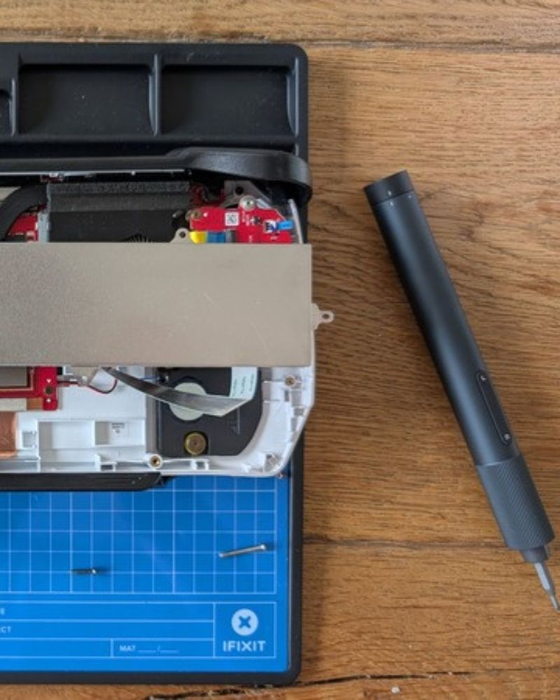 A partially ROG Ally on an iFixit mat with a black screwdriver nearby, showing internal components like a fan and circuit board.