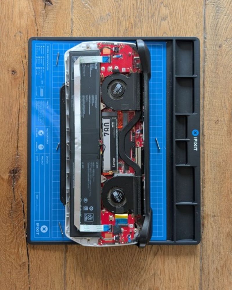 ROG Ally internals on a repair mat showing internal components, including a visible JSAUX battery, dual cooling fans, and circuit board.