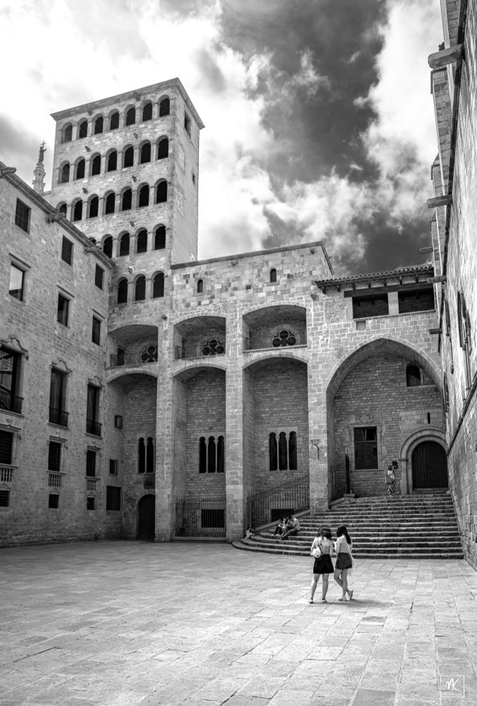Black and white photo of a courtyard surrounded by stone buildings with two people standing in it. 