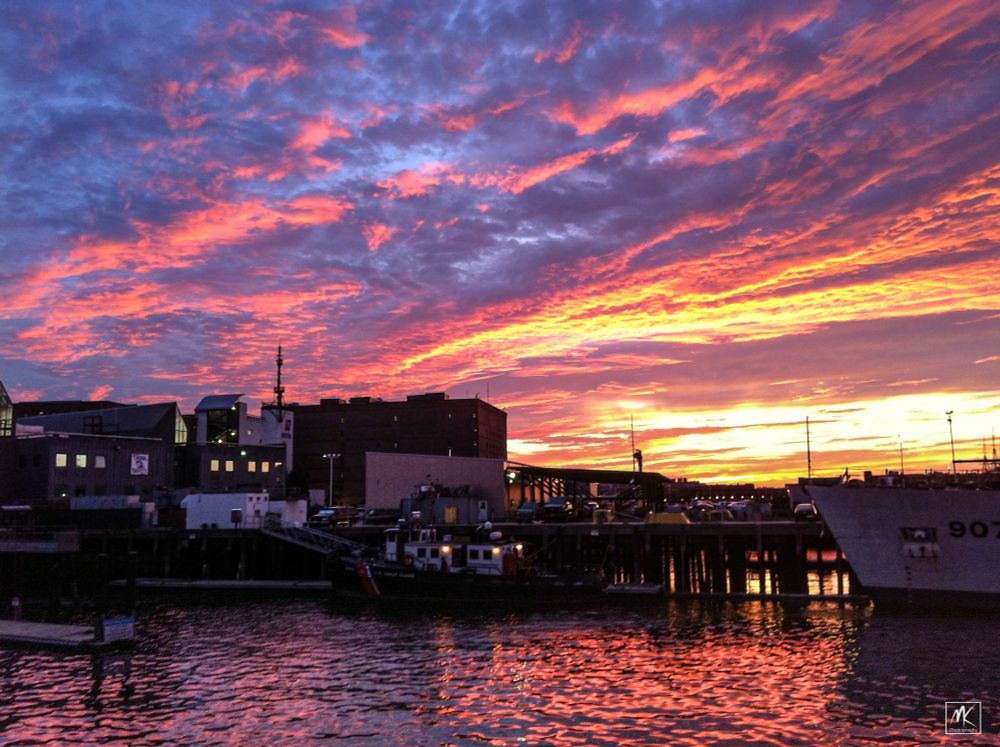 Color photo of a very colorful sunset sky over a waterfront scene with the sky’s colors reflected in the water in the foreground.