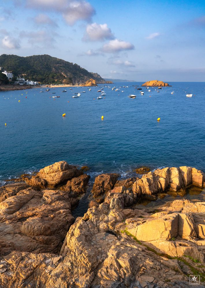 Color photo taken late in the afternoon with a rocky shoreline in the foreground, a small bay with moored boats in the middle ground, and a hilly coastline stretching off into the background distance. 