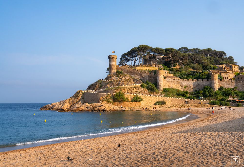 Color photo with a sandy beach lot by the late afternoon sun in the foreground and beyond it is a winding path up a seaside hill that is surrounded by medieval walls and towers.