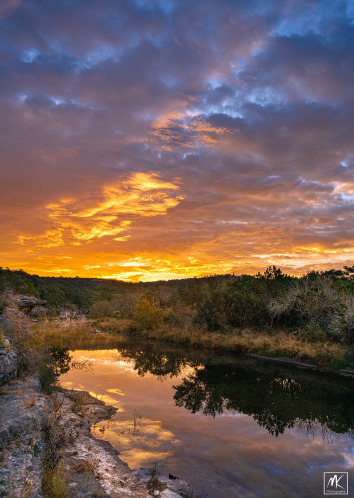 Color photo of a colorful autumn dawn sky over the still waters of a creek in the Texas Hill Country.