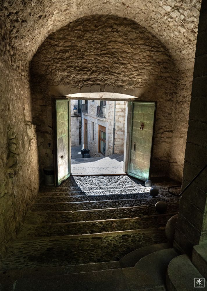 Color photo taken looking down stone steps in a barrel-vaulted space through an open doorway at the end of it with a street and buildings beyond. 