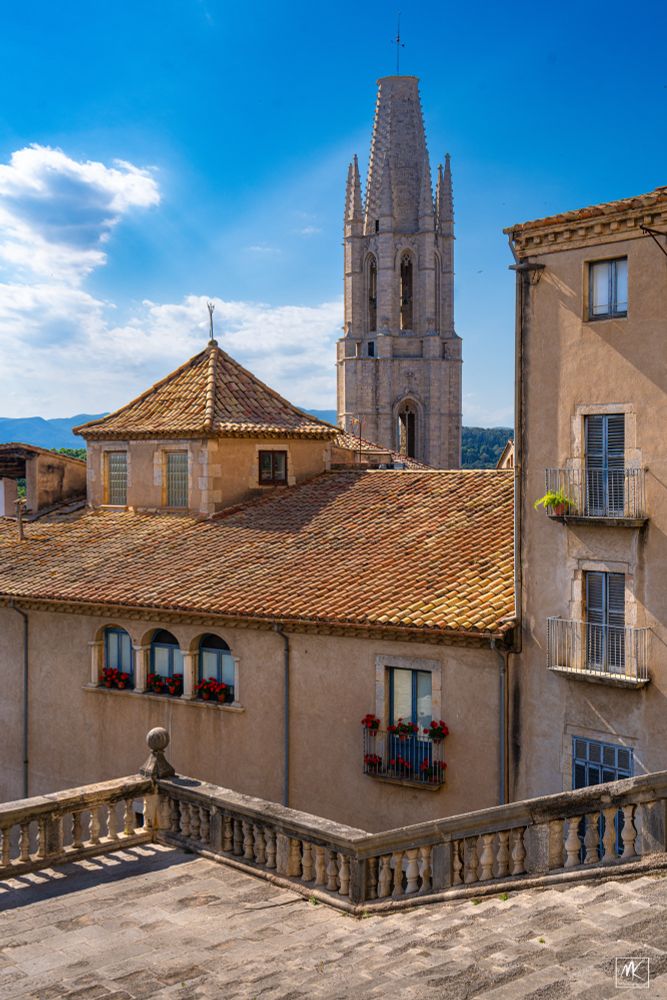 Color photo of a city scene with broad steps in the foreground and tile roofed buildings beyond them and a tower in the background. 