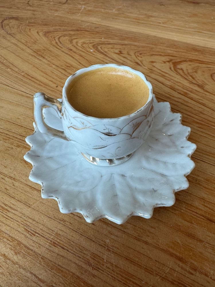 Ornate debutante tea cup, white with gold inlay sitting on a matching saucer with a lead pattern and points at the edges like a flower. The cup has espresso inside and the saucer rests on a wooden table.