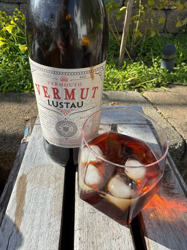There is a bottle of vermouth “Vermut Lustau” and a glass with said vermouth, ice and some soda water sitting on a faded wooden table. In the background is a stone wall just an inch or so shorter than the table with a raised garden bed behind it filled mostly with weeds and a small blueberry bush. Behind the bush is another stone wall.

The vermouth is quite delicious and has a complex and fragrant aroma of wormwood, gentian, coriander and orange peel. Highly recommend.