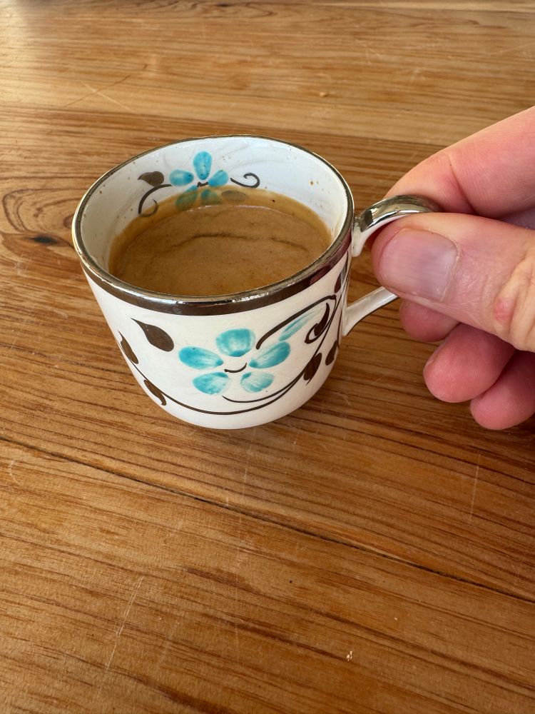 Hand holding a debutante tea cup filled with espresso. The cup is shaped like a tiny coffee mug but ornately decorated with a silver and light blue flower pattern. In the background is a wooden table.

This cup is tiny
But the flavor bold
Easy to drink, hard to hold