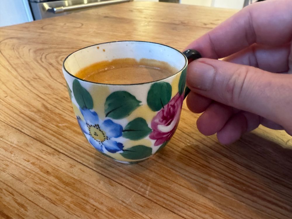 Yet another debutante tea cup is held by my hand over a wooden table. The cup is decorated with colorful blue and red flowers with green leaves and splotches of yellow. 

