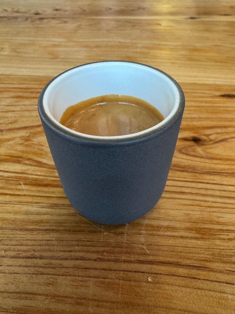 A grey ceramic espresso cup with a white interior and espresso inside sitting on a wooden table. 

The coffee calls to me
Beckoning me to drink an embrace the day
A the bottom awaits a moment of clarity 
And the need to poop.