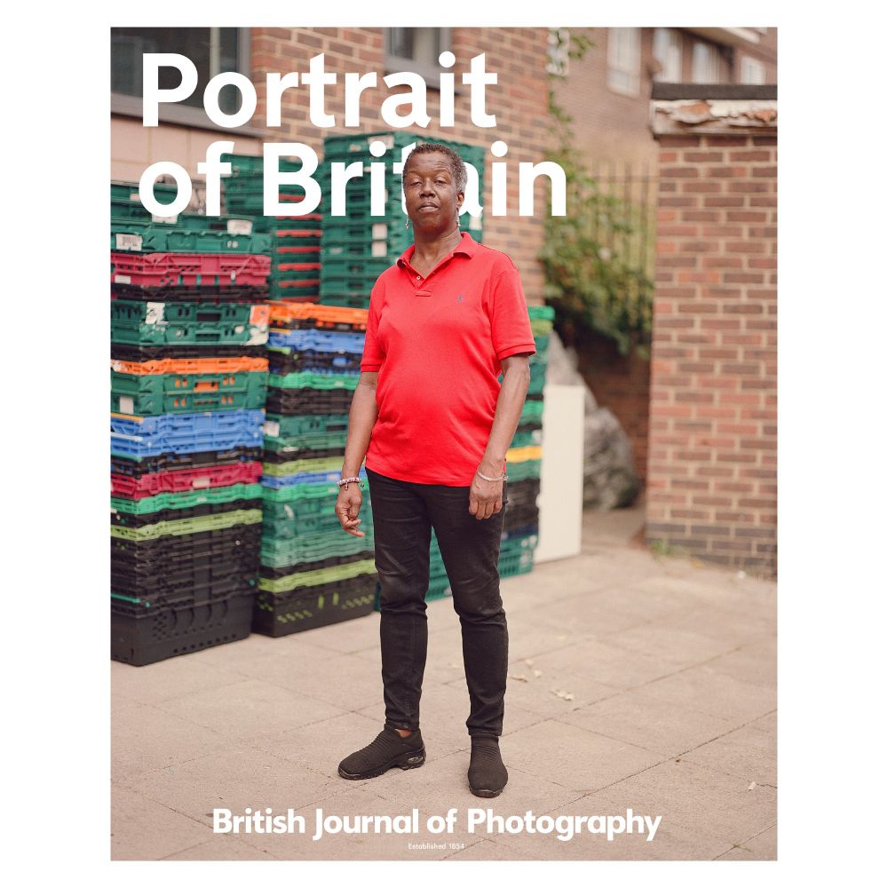 Dawn, a foodbank volunteer, stands in front of crates stacked up behind her. She is working on supply deliveries, carrying them into the centre to be sorted. She is wearing a red top, black trousers and black trainers. Behind her are the words 'Portrait of Britain'. Featured in the British Journal of Photography. 