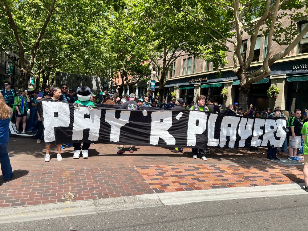 Sammy Sounder holding a banner that reads “PAY R PLAYERS” with the Emerald City Supporters in support of the Sounders players and MLS Players Union.