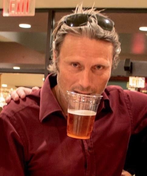Mads Mikkelsen holding a cup of beer with his teeth