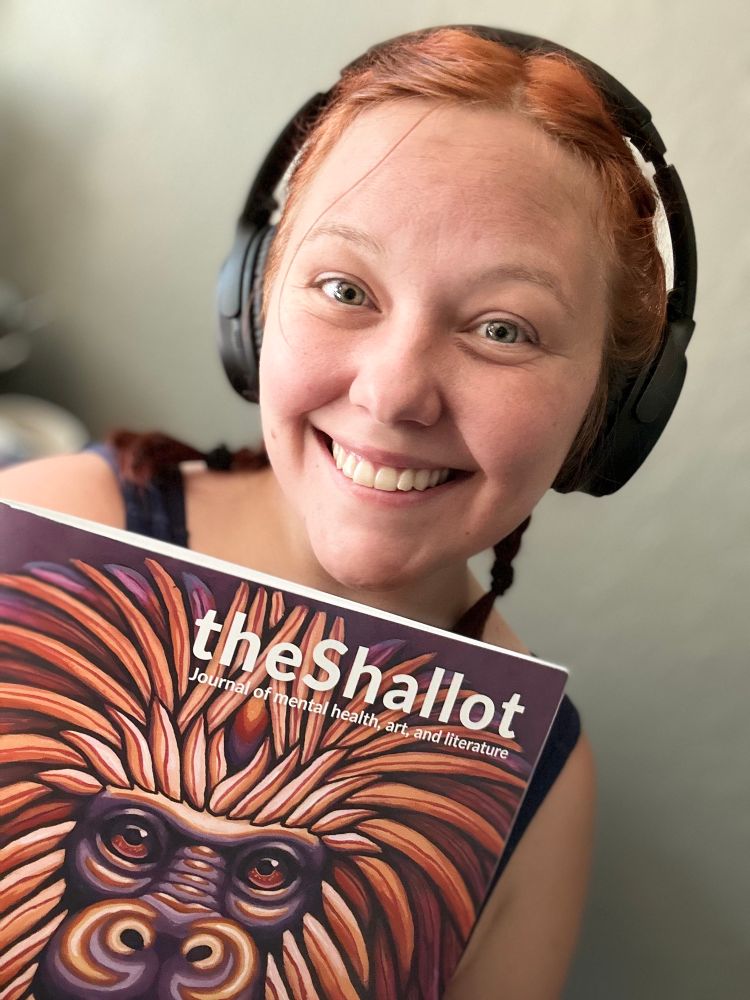 Photo of the artist (fair skin, red auburn hair) smiling, holding a magazine “The Shallot: Journal of mental health, art, and literature” with a painted monkey on the cover