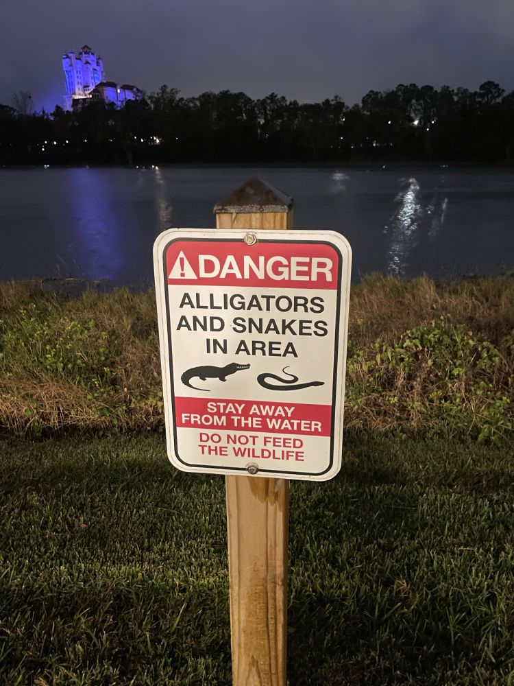 Sign in front of a lake saying “Danger. Alligators and snakes in area. Stay away from the water. Do not feed the wildlife.” The sign is white with red and black letter and has a drawing of an alligator and a snake