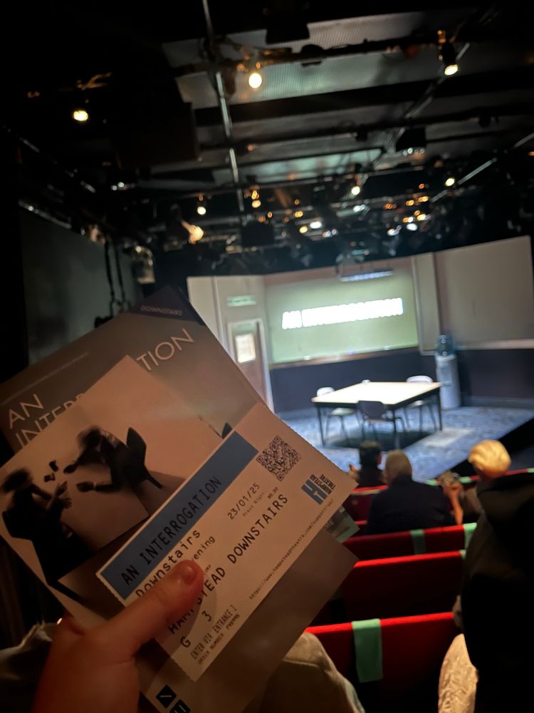 A ticket, playtext, and programme for the show ‘An Interrogation’ is clutched in the foreground, with red seats, a rectangular table, water cooler, and three black chairs, with a projection illuminating ’An Interrogation’, can be seen in the background.