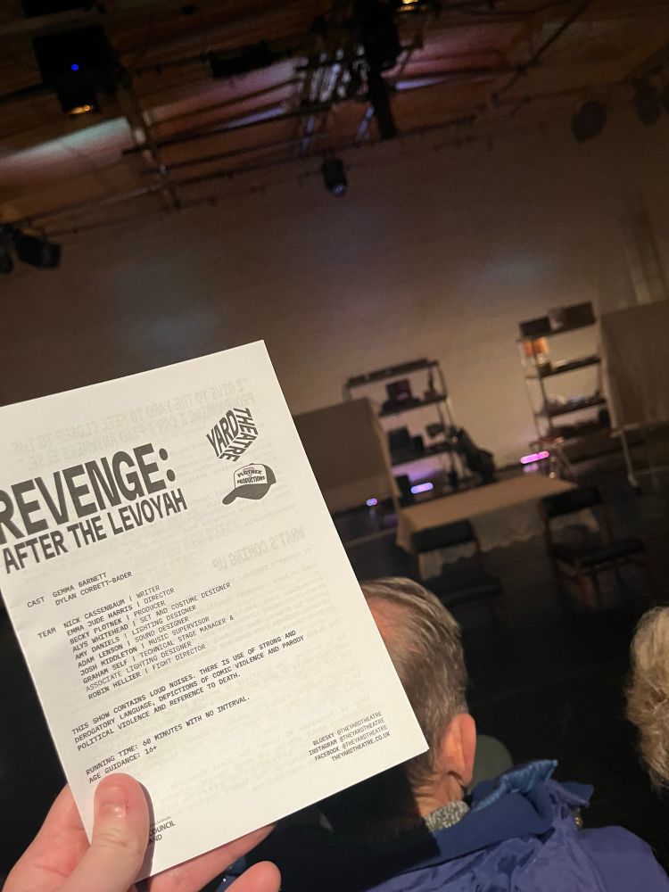 A free sheet for the show, Revenge: After the Levoyah, in the foreground, with the set beyond that, made up of a table and two chairs, shelving units, and white boards covered in fabric.