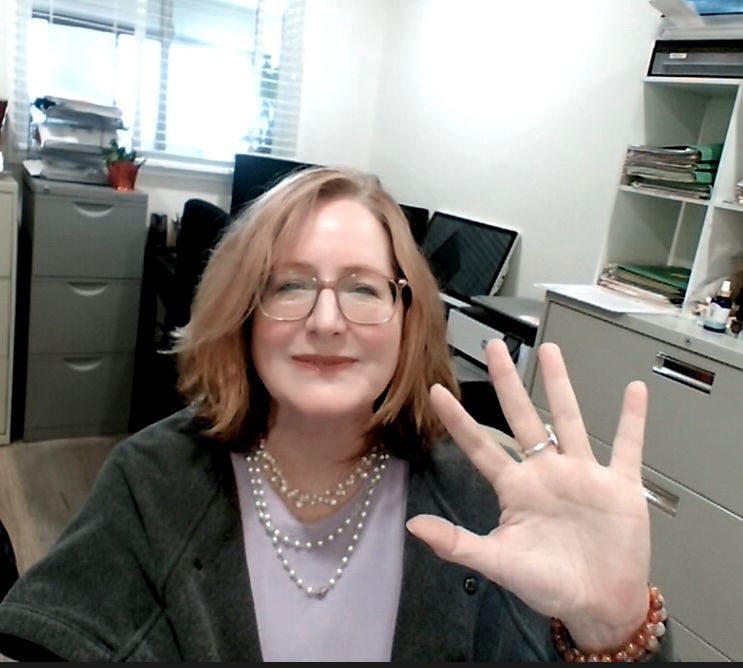 Glasses wearing cishet white lady in a lavender top and grey sweater wearing a beaded necklace waving at the camera 