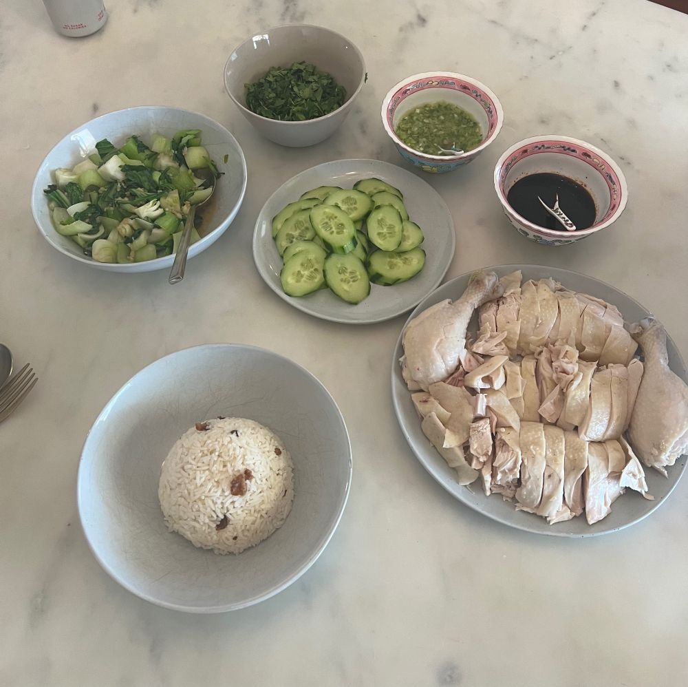 Homemade Hainan chicken rice with rice in foreground, poached chicken on right and stir fried bok choy and cucumbers with associated sauces behind 