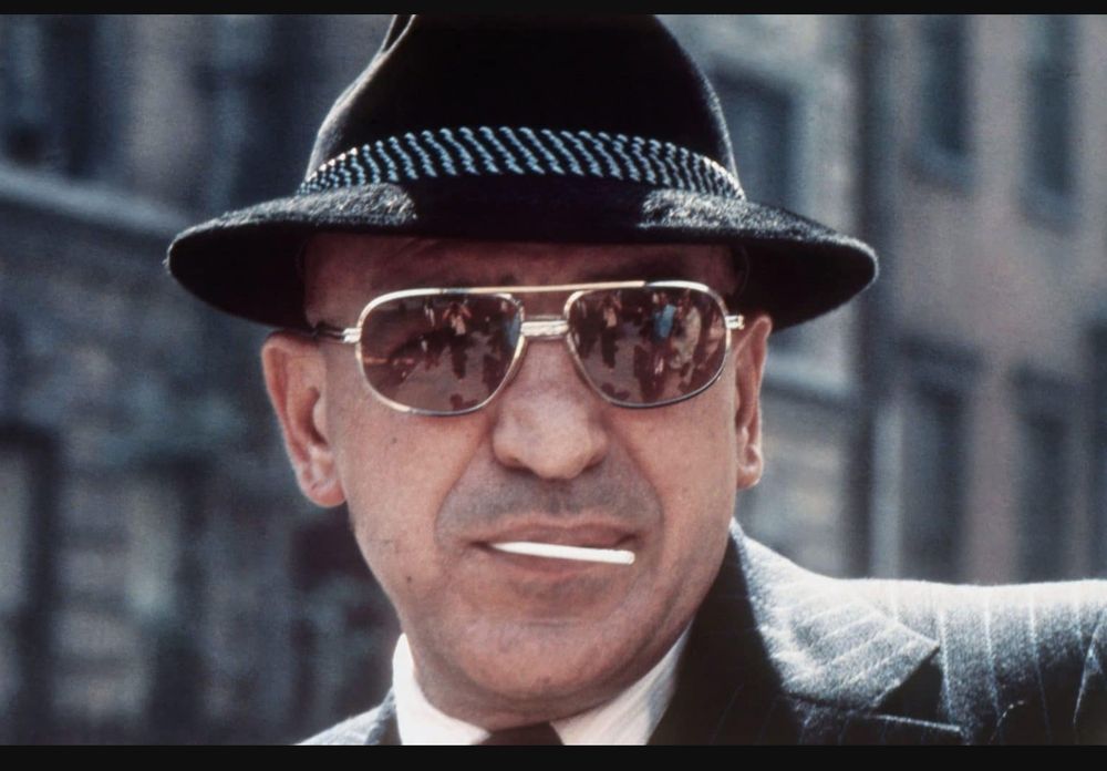 Facial photo of man wearing fedora, striped suit jacket and sunglasses with a lollipop stick in his mouth 