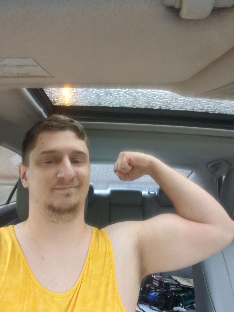 White man sitting in a car flexes his bicep. He's wearing a yellow tank top and smirking