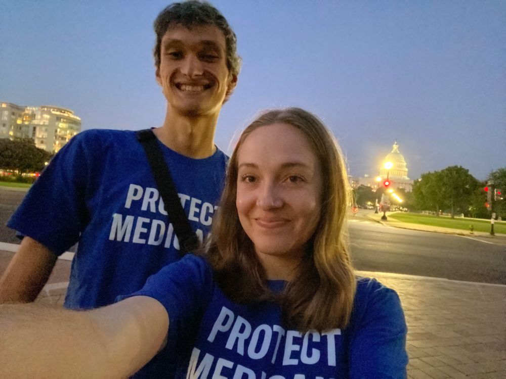 Selfie of Casey, a white woman with blonde hair, and Charles, a multiracial man with black hair, in front of the US Capitol building at dusk. They are both wearing blue shirts that say "Protect Medicaid."