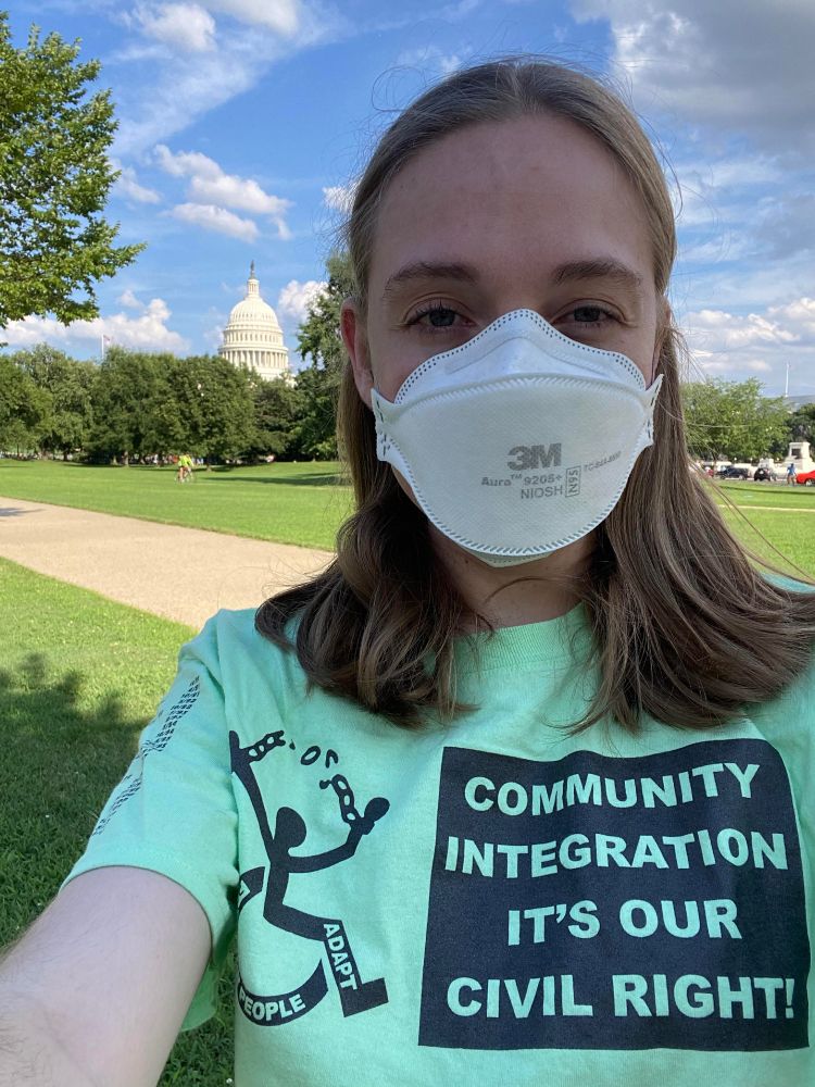 Selfie of Casey, a white woman with dark blonde hair wearing a white 3m Aura N95 mask, with the US Capitol building in the distance. Casey is wearing a green shirt that depicts a wheelchair user breaking free of chains and says “Community Integration: It’s Our Civil Right!”