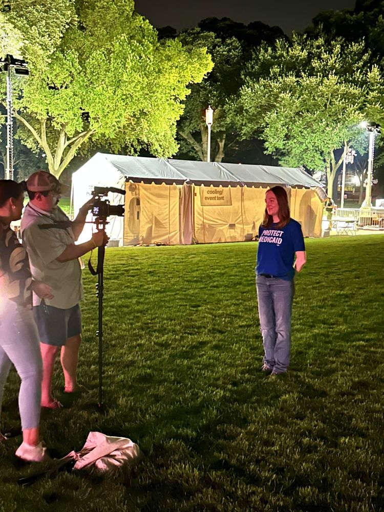 Casey stands in front of a reporter and a videographer. They are outside at the Medicaid Vigil at night, in front of a white cooling/event tent and trees.