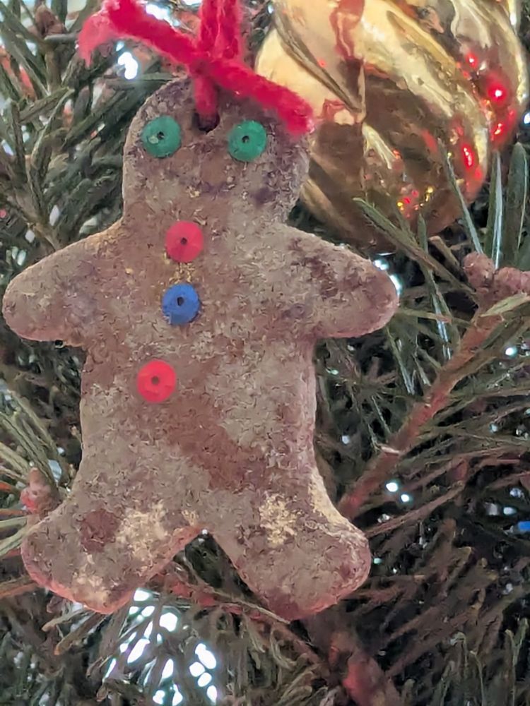 An old gingerbread man ornament with a red ribbon and sequin eyes and buttons of various colors. Handing on a Christmas tree 