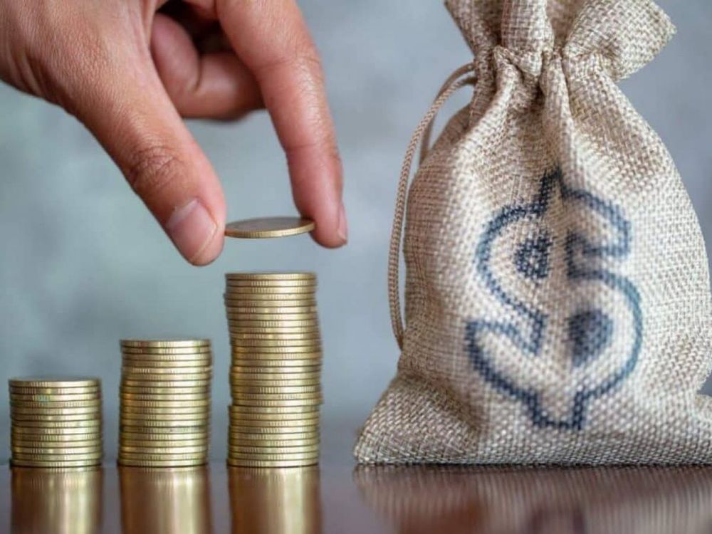 Mão empilhando moedas douradas. Ao lado um saco com um cifrão desenhado