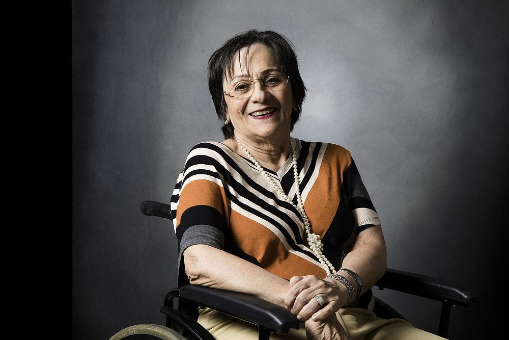 Imagem mostra Maria da Penha uma mulher idosa sentada em uma cadeira de rodas, sorrindo para a câmera. Ela usa óculos, tem cabelos curtos e escuros, e veste uma blusa com estampa geométrica em tons de preto, branco e laranja. Também usa um colar de pérolas e pulseiras, transmitindo uma imagem elegante e simpática. O fundo é neutro e escuro, contrastando com a iluminação suave sobre seu rosto.
