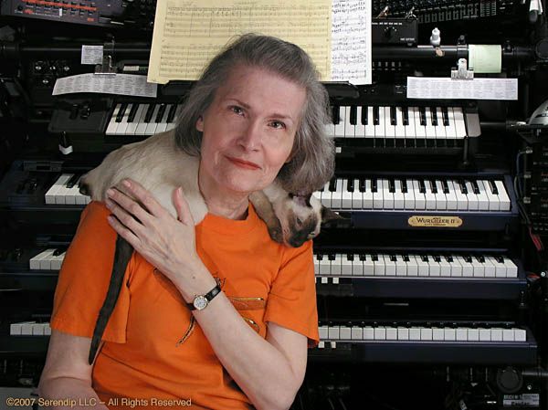 Wendy Carlos sitting in front of a bunch of synthesizers with a cat resting on her shoulders