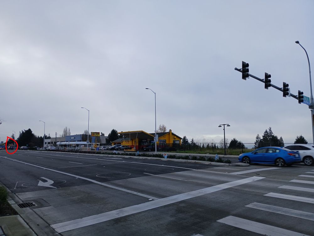 Crosswalk on a seven lane road with a tiny red circle on the left highlighting a bus stop