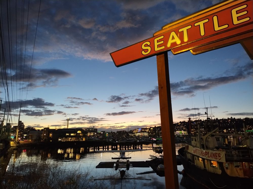 Lake Union with docked seaplane at dusk, Space Needle in the distance with swirling clouds overhead. A "Seattle" sign dominates the foreground 
