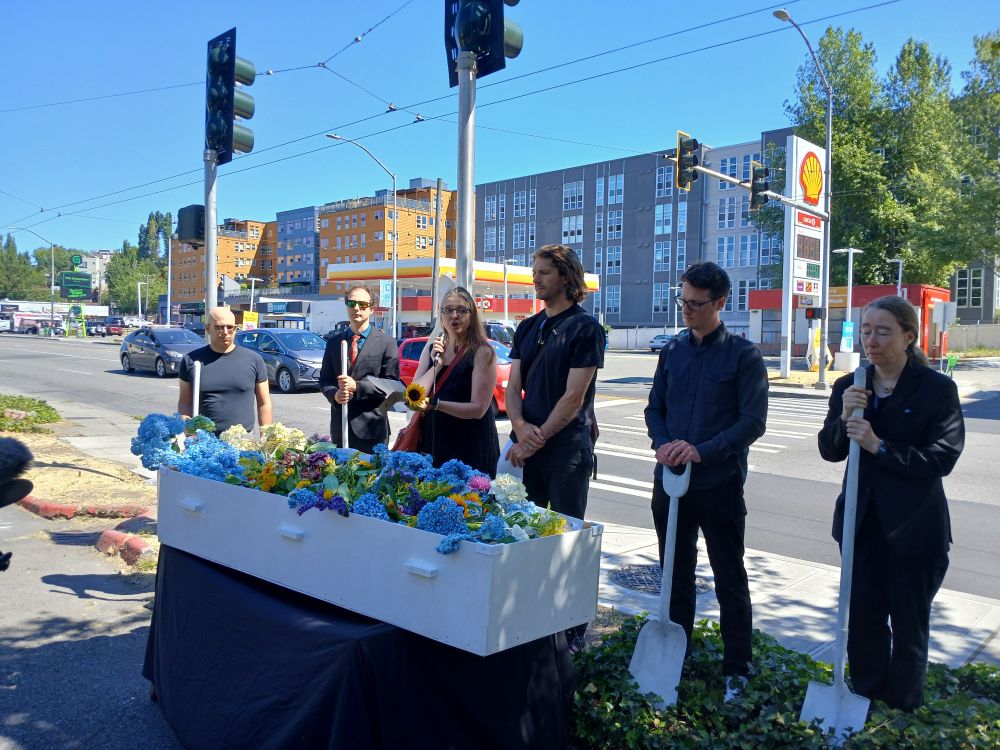 Folks gathered around a white coffin filled with flowers, holding while shovels