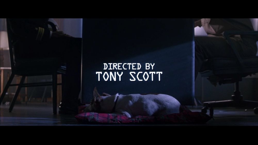 2 persons sitting at a desk. We don’t see the heads. 
A dog is sleeping on a dog bed in the middle. 

Text in the middle of the screen reads « directed by Tony Scott »