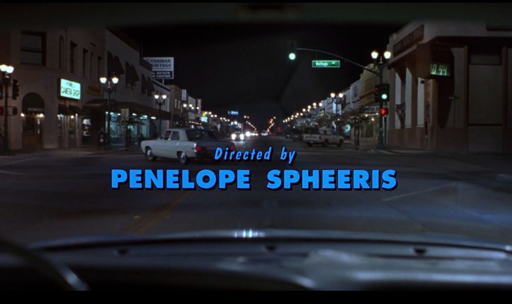 Credits from Wayne’s World (1992)
POV from a car. A street. With cars. 

Text in blue : “Directed by Penelope Spheeris“