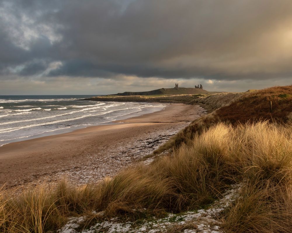 A photo of a beach with sand dunes behind. In the foreground dune grasses are caught in sunlight as the sun breaks through cloud to the right of the frame. In the distance a ruined castle stands on top of a small hill. The sky is cloudy.