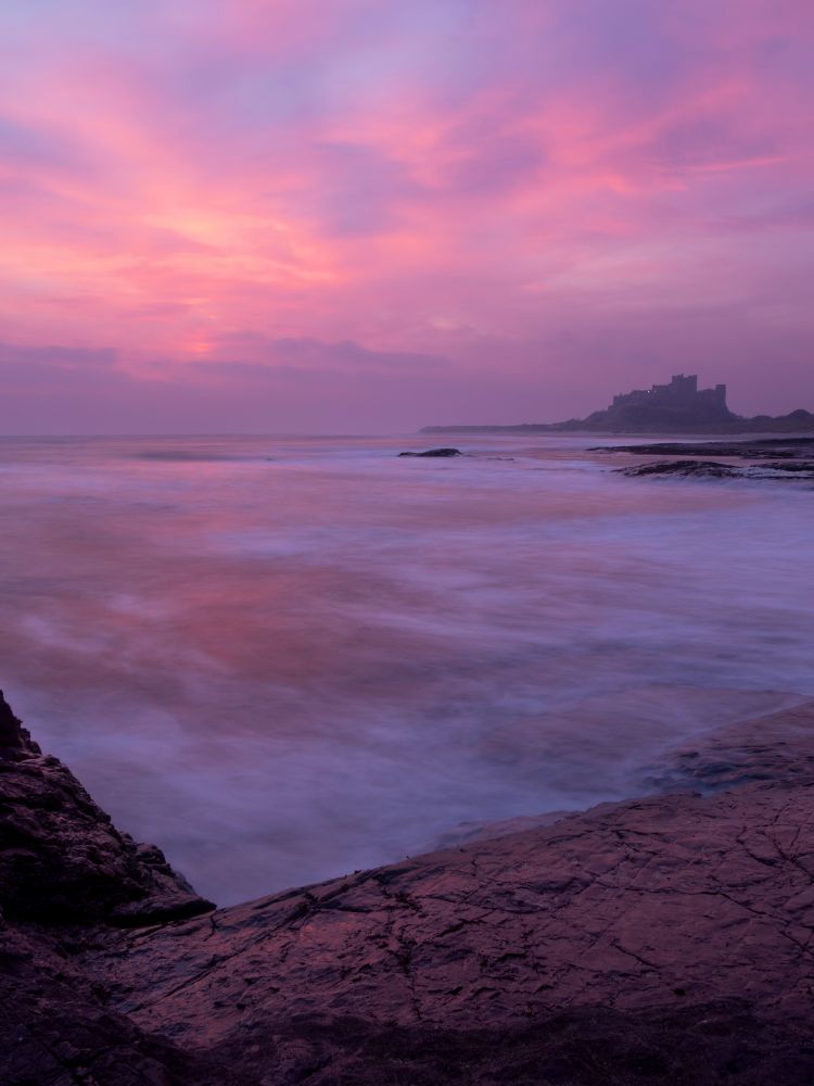 A photo taken at sunrise. On the horizon stands Bamburgh castle silhouetted against a partly cloudy coloured pink sky. 

In the foreground are rocks, which reflect back the pink hues of the sunrise. In the mid ground the sea has been frozen and smoothed out with a long exposure, the pinks of the sky reflected back towards the camera in the calmer parts. Other rougher parts are white with a misty appearance. 
