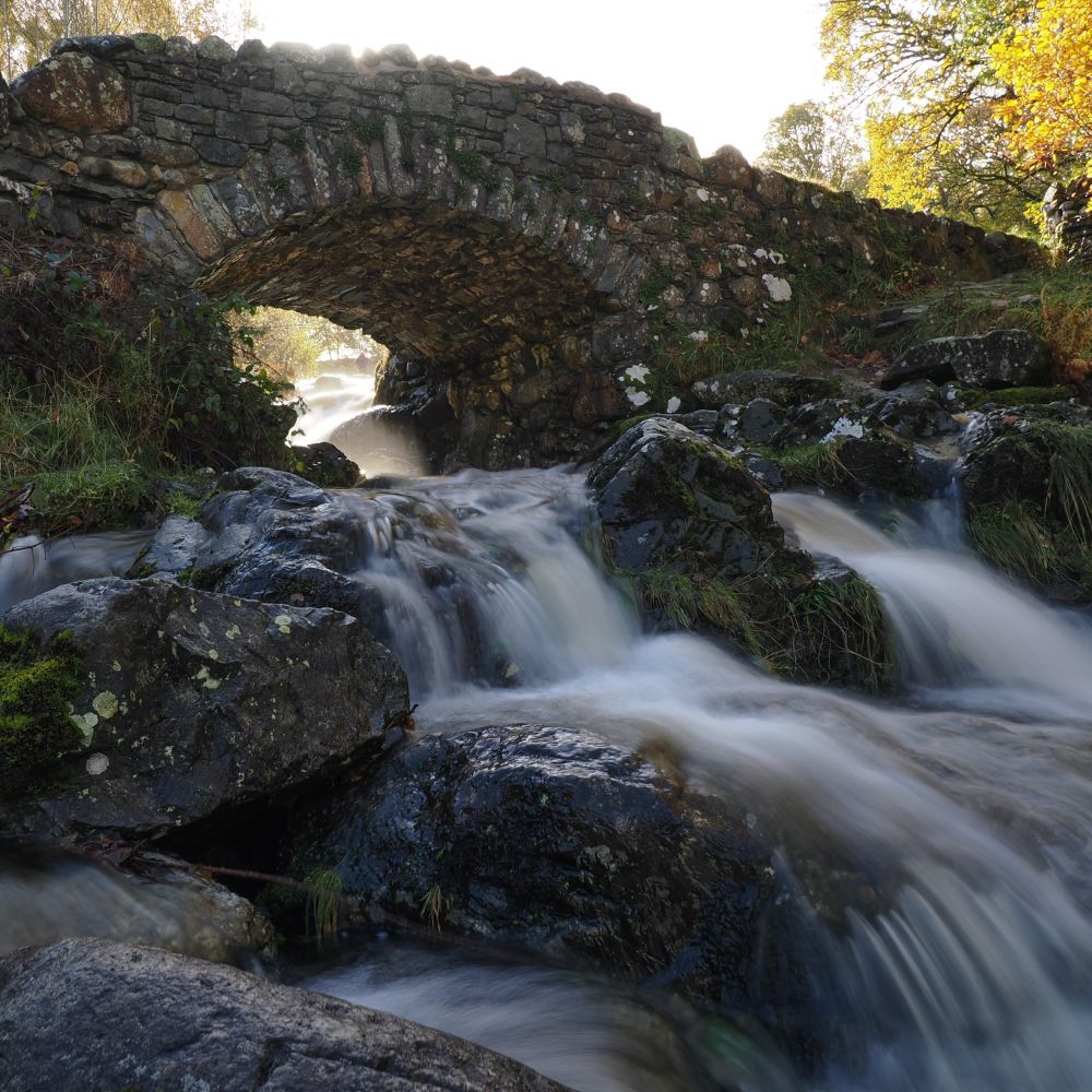 In the foreground a stream tumbles over rocks, the movement on the water captured with a long exposure. Above the stream a stone arch bridge. 