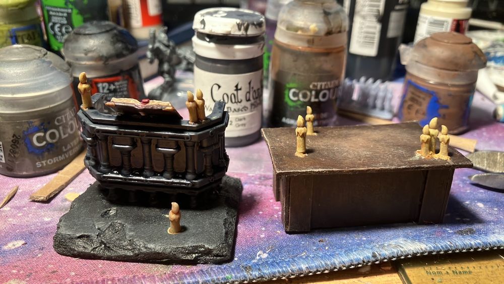 two more pieces of tiny church furniture. the first is mantic games’ holy tome terrain piece (a plastic 28mm-ish scale lectern with a book on it) attached to a piece of foam board as a base that is weathered to look like dark grey stone. the lectern is painted a cool grey, and not yet highlighted (though my lamp is doing it’s best to make it look like it is). I have added 3d printed candles to it and the “stone” in front. they are currently painted warm beige, including the flames, because I haven’t finished them yet. next to it is a high table (maybe a bit too high, scale-wise) built out of card and wooden drink stirrers, and painted in the same style as the pews and kneelers. there are clusters of 3d printed candles on it, too, also unfinished and beige. 