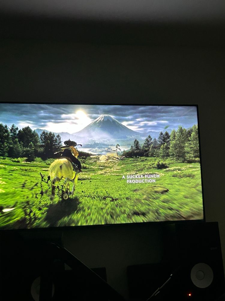 Photo of a Ghost of Yōtei video game on a tv, animated woman samurai on horseback in a field of green with mountains, trees and sun glaring in the distance 