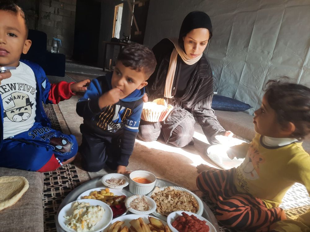 A Palestinian woman sits in the background in a concrete room partially wallpapered with plastic sheeting, as two small boys and a girl enjoy treats from a small tray of foods and dips.