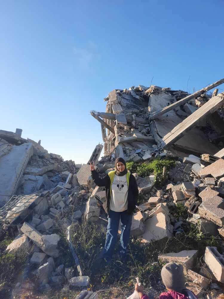 A Palestinian woman wearing baggy clothing stands amid concrete ruins under a blue sky. At the very bottom of the image, a small child is waving to her.