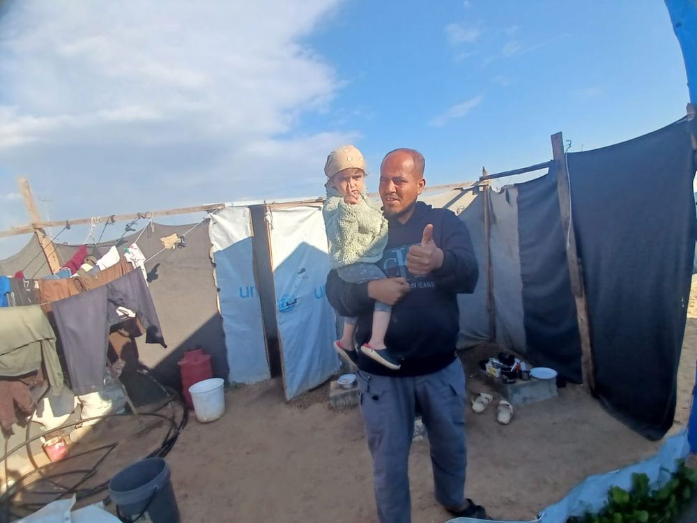 A Palestinian man holds a three-year-old Palestinian girl and they both make thumbs-up signals as they stand in a small, sandy courtyard enclosed by plastic sheeting, with laundry drying at one side.