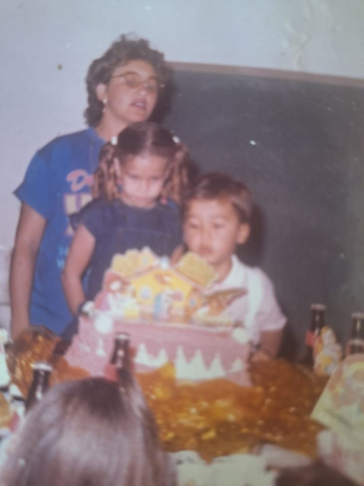 Minha mãe usa óculos de grau e camiseta azul, eu uso vestido jeans e tenho cachinhos no cabelo que é partido no meu e preso com duas chiquinhas. Meu irmão usa camisa branca e vermelha e macaquinho branco. Na nossa frente tem um bolo enorme de glacê branco e rosa e atrás de nós um quadro negro de escola 