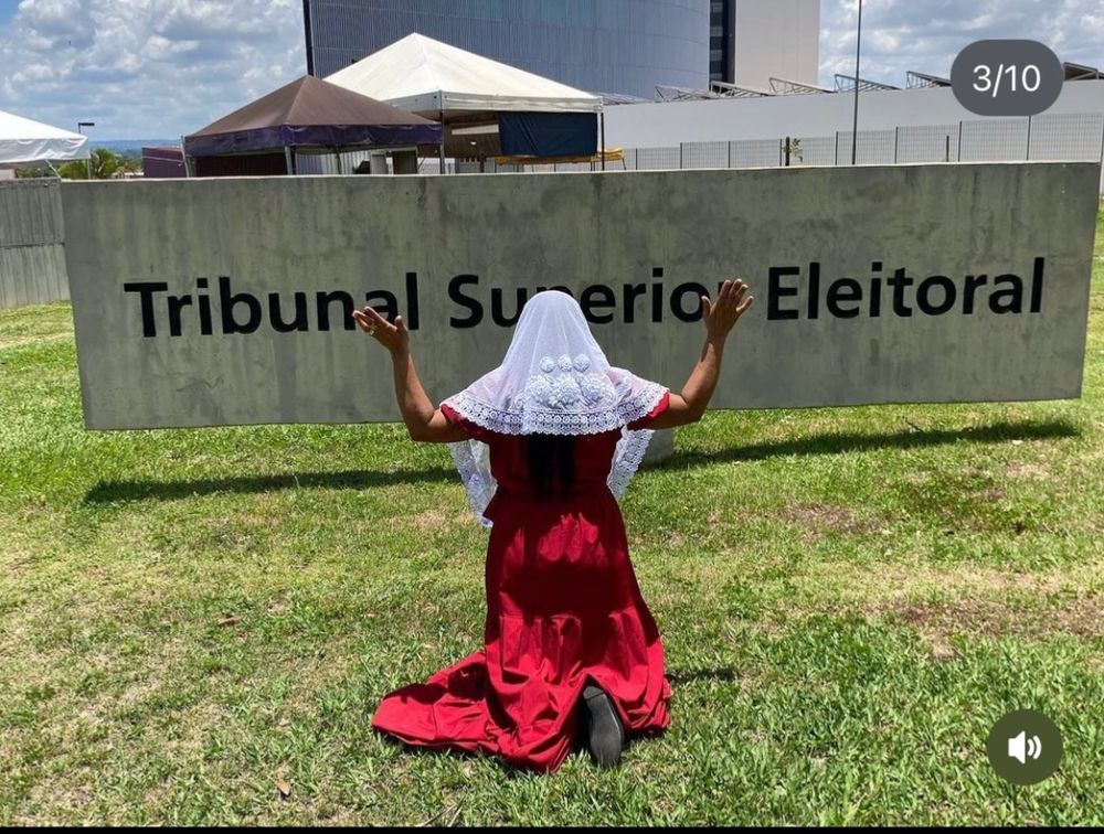 Uma mulher de vestido vermelho e véu branco está de joelhos e braços levantados em posição de oração e benção em frente ao tribunal superior eleitoral