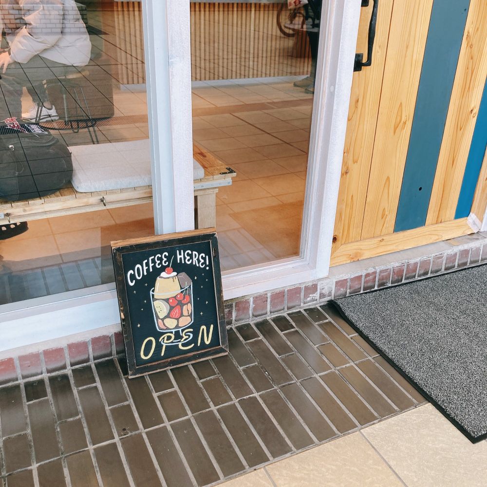 店の入口下部と外の床タイルを店の外側から切り取って写した写真。

奥の店はガラス張りで店内の様子が少しだけ見え、入口ドアは明るい木目調で一部ブルーグリーンに塗装されている。

手前のタイルは店に近い部分が焦茶色で、通路側がベージュ。店のドアの前に灰色のマットが敷かれている。

タイルの上に、店の外壁にもたれるように小さな看板が置かれている。プリンアラモードのようなイラストがカラーで描かれ、COFFEE HERE! OPENとある。